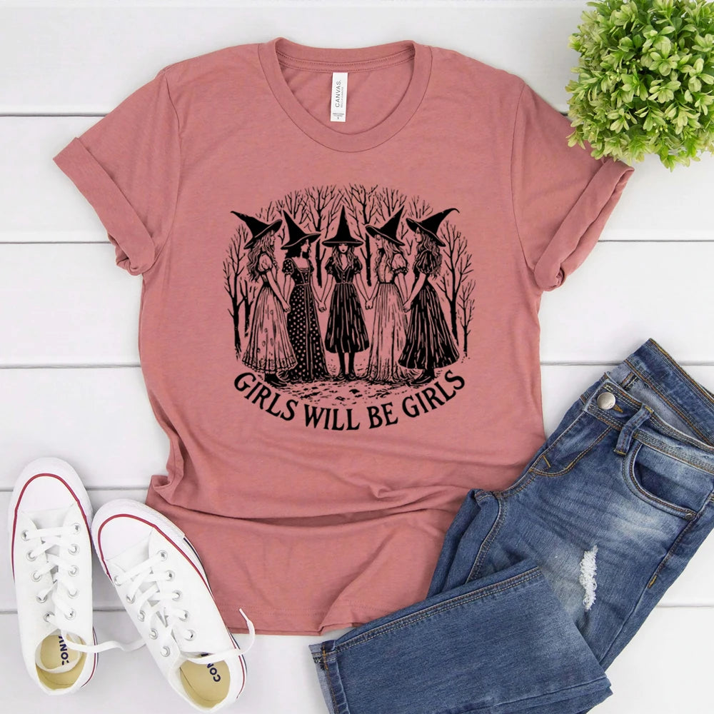 Pink t-shirt with witch graphic and 'Girls Will Be Girls' text, paired with jeans and sneakers on a white wooden surface.