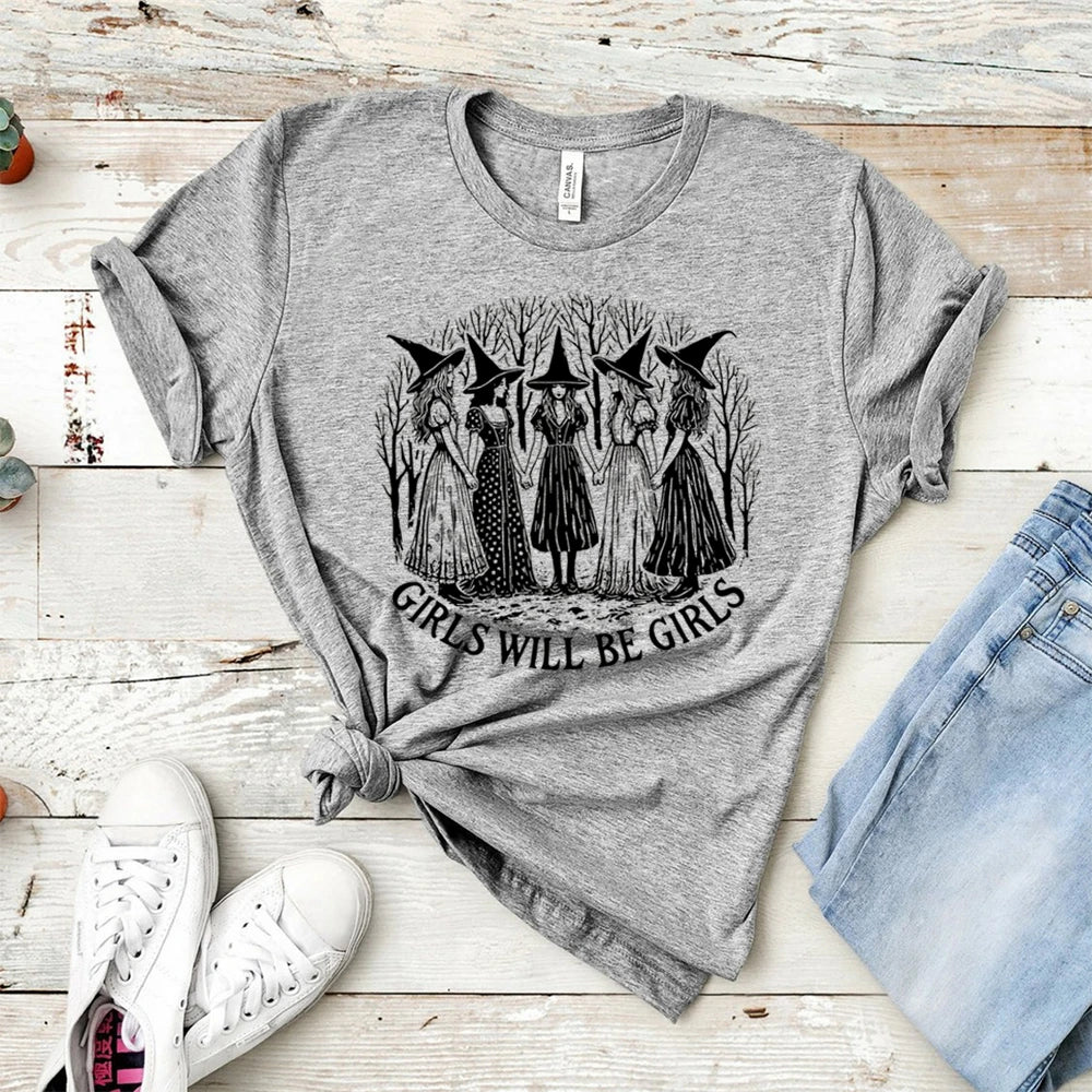 Gray t-shirt with witch graphic and 'Girls Will Be Girls' text on a wooden surface with jeans and sneakers.